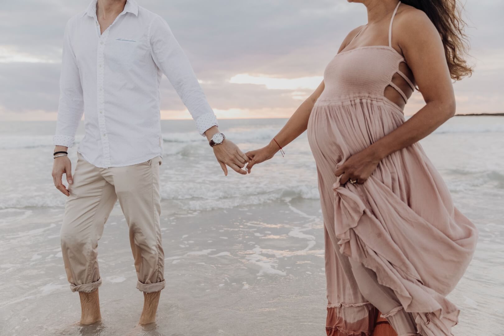 Exuberant pregnant woman wearing a flowy sundress and her supportive husband standing hand-in-hand on the white sandy beach of Miami, Florida, gazing out onto the vast expanse of the bright blue ocean.