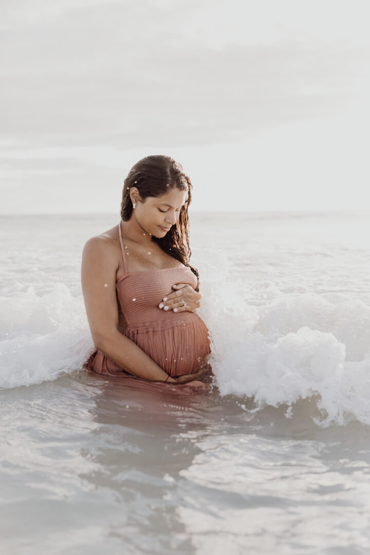 The silhouette of an ecstatic pregnant woman stands before an ethereal backdrop of orange-red skies on Miami beach, cradling her blooming belly adorned by a wet sundress.