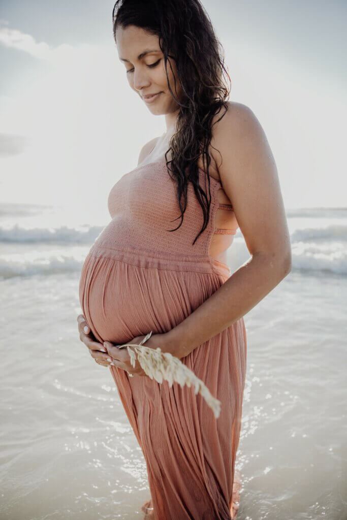 A pregnant woman in a sundress stands facing to the left, hands covering her pregnant belly, with a stunning sunrise in the background at Miami Beach.